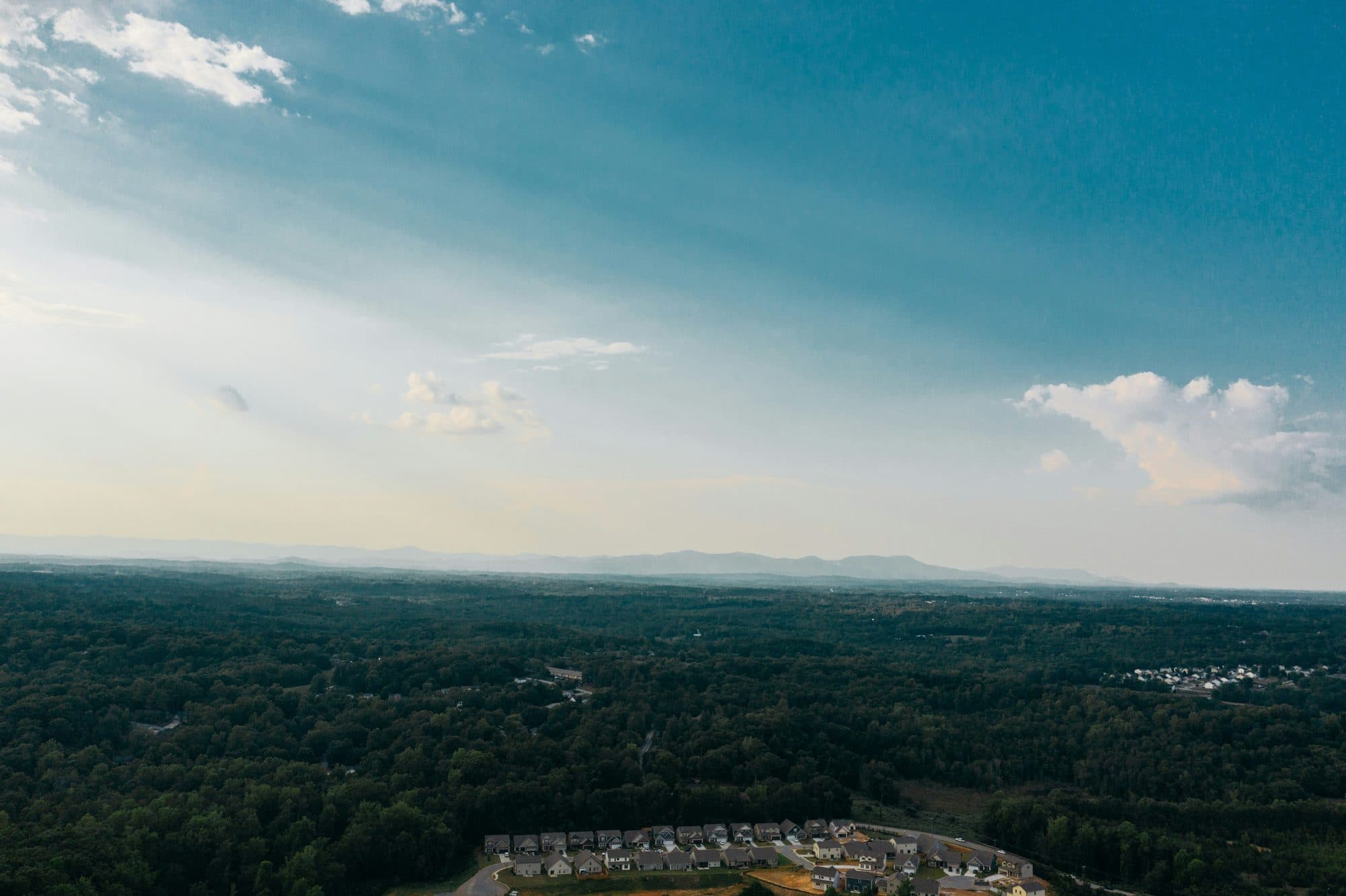 Aerial view over wooded land near Taylors, SC, with sunbeams in a blue sky and mountains on the horizon.
