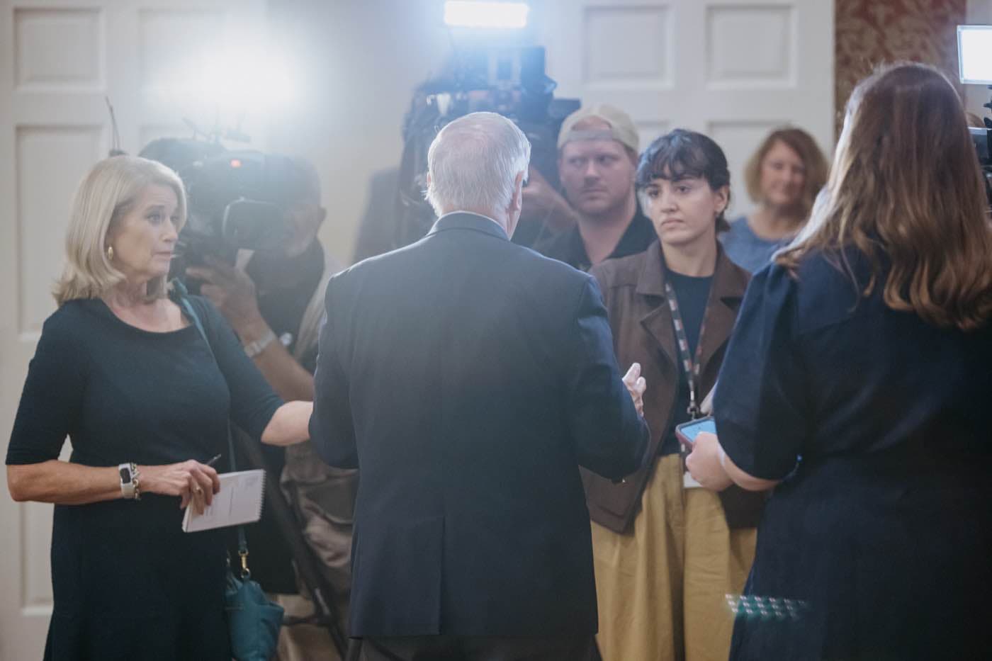 A man in a suit speaks to a group of reporters and camera operators in a brightly lit room.