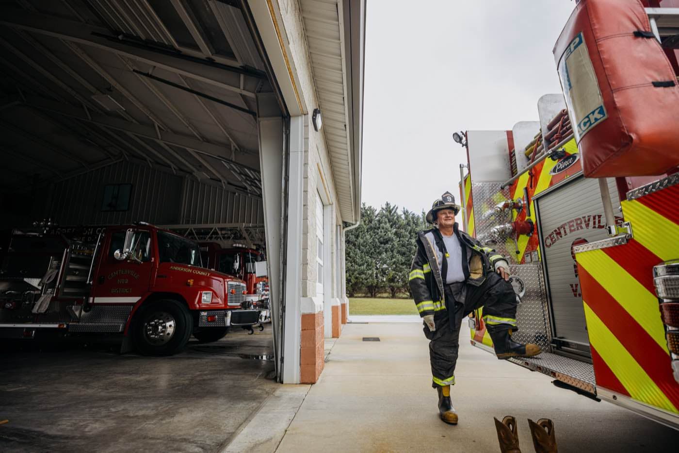 Firefighter in full gear, one foot on a fire truck, looking at the camera with a smile.