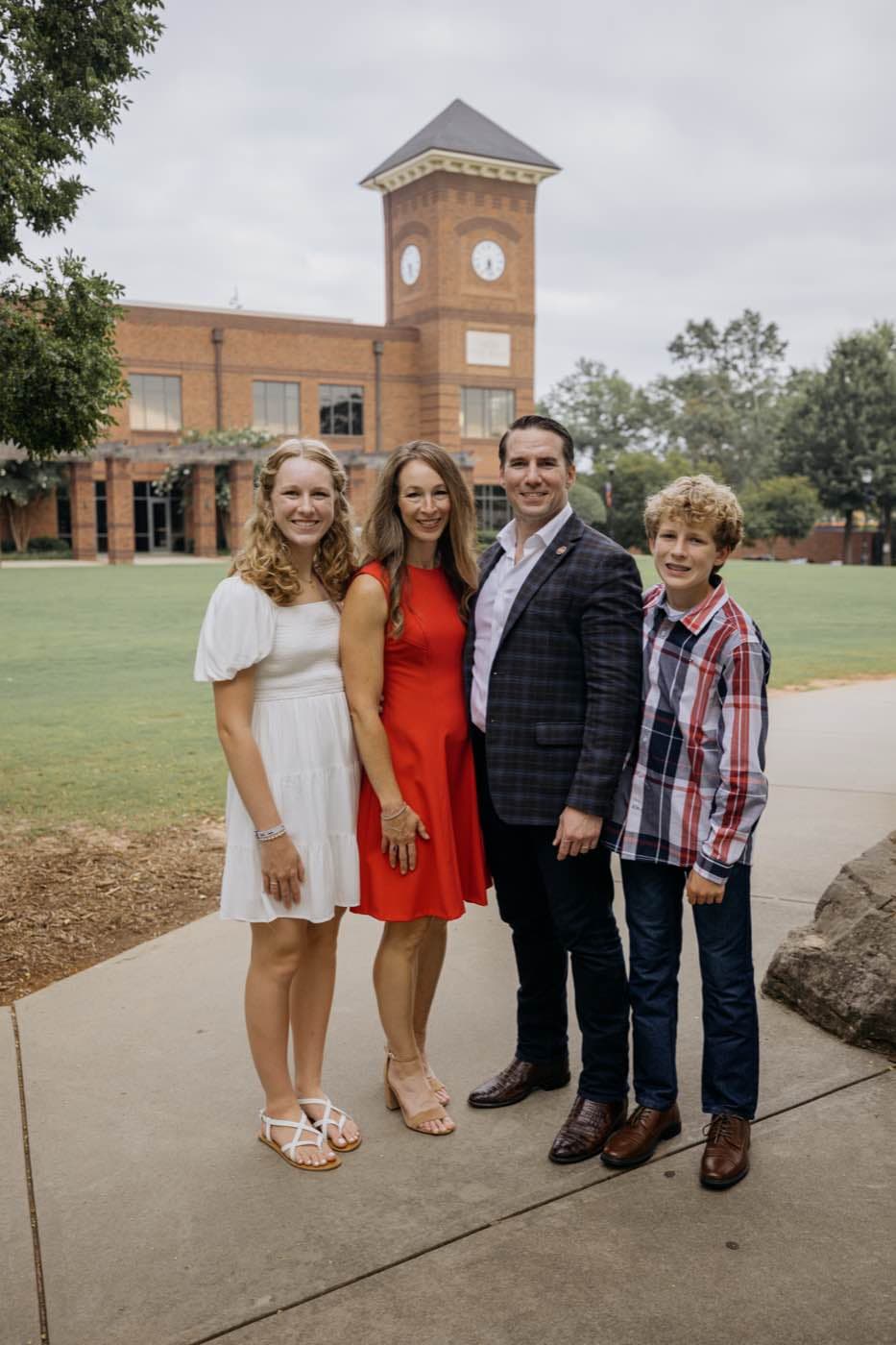 A family of four smiling outdoors in front of a brick building with a clock tower on a cloudy day.